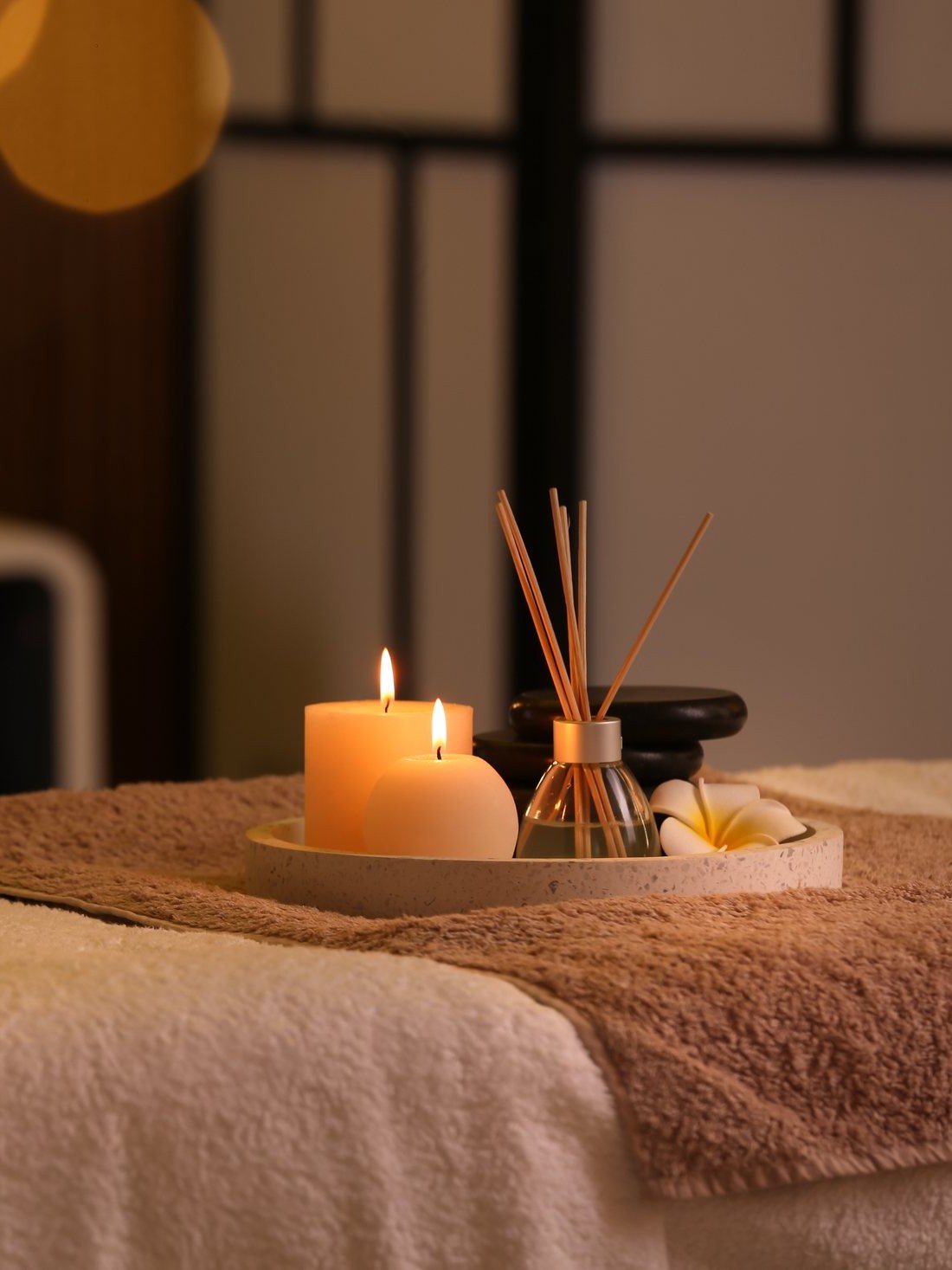Lit candles, a reed diffuser and smooth black stones on a tray, resting on soft towels.